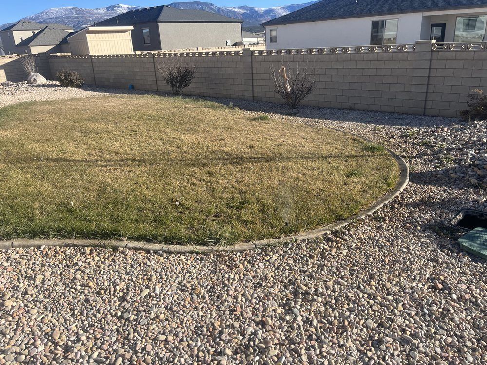 Backyard with brown/green grass, rock border, rock ground, beige block wall, and distant snow-capped mountains.