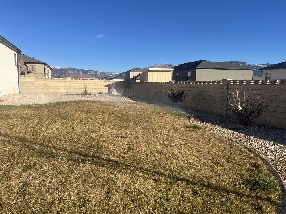 Sprinkler watering a dry, brown lawn in a backyard; tan brick wall and houses in the background.