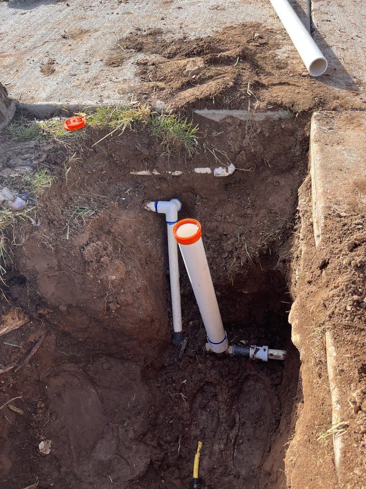 Excavated ground revealing PVC pipes, fittings, and an orange-capped vertical pipe, near a concrete edge.