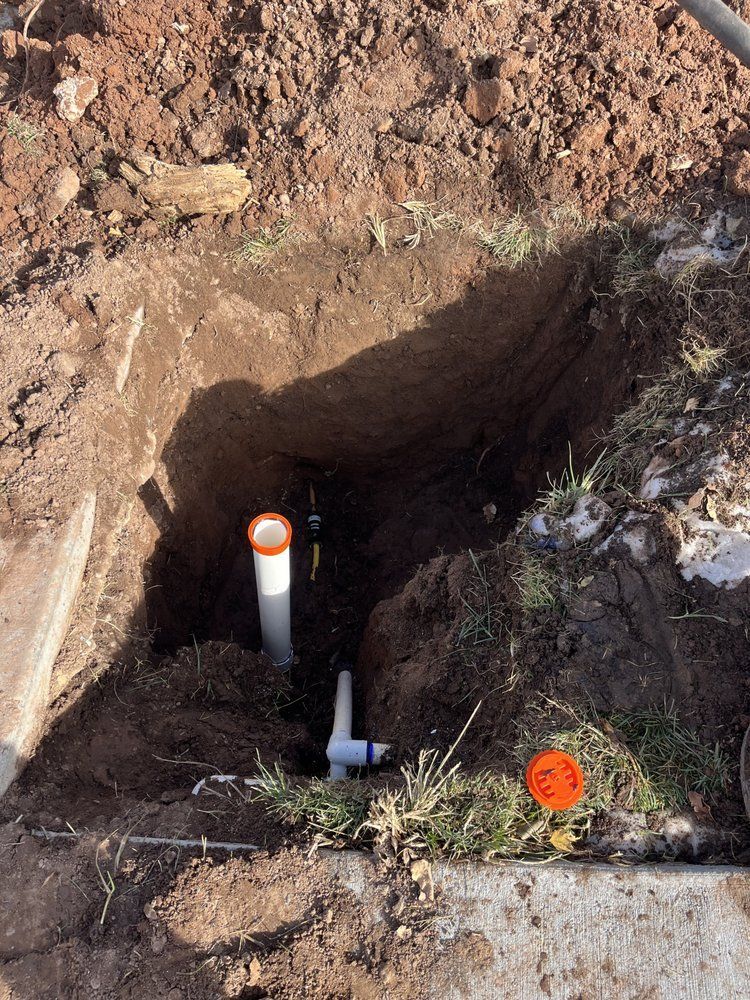 Open hole revealing white PVC pipes and orange caps buried in dirt.