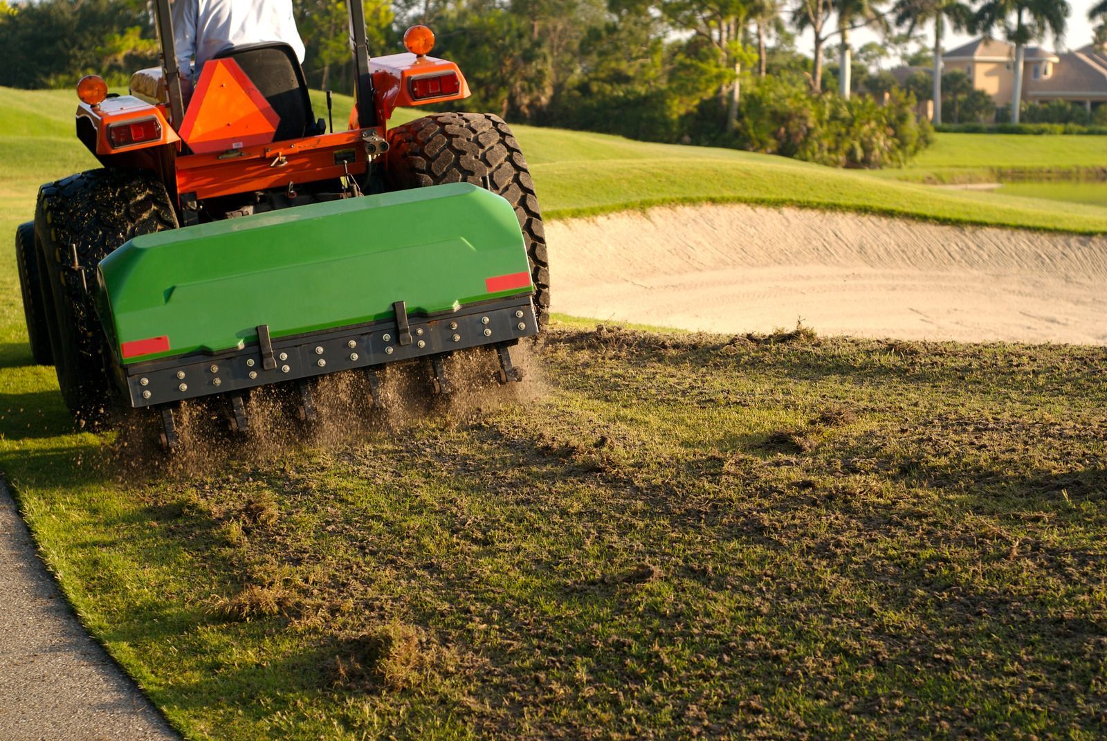 Tractor aerating a golf course green, with green and brown grass, and sand bunker in the background.
