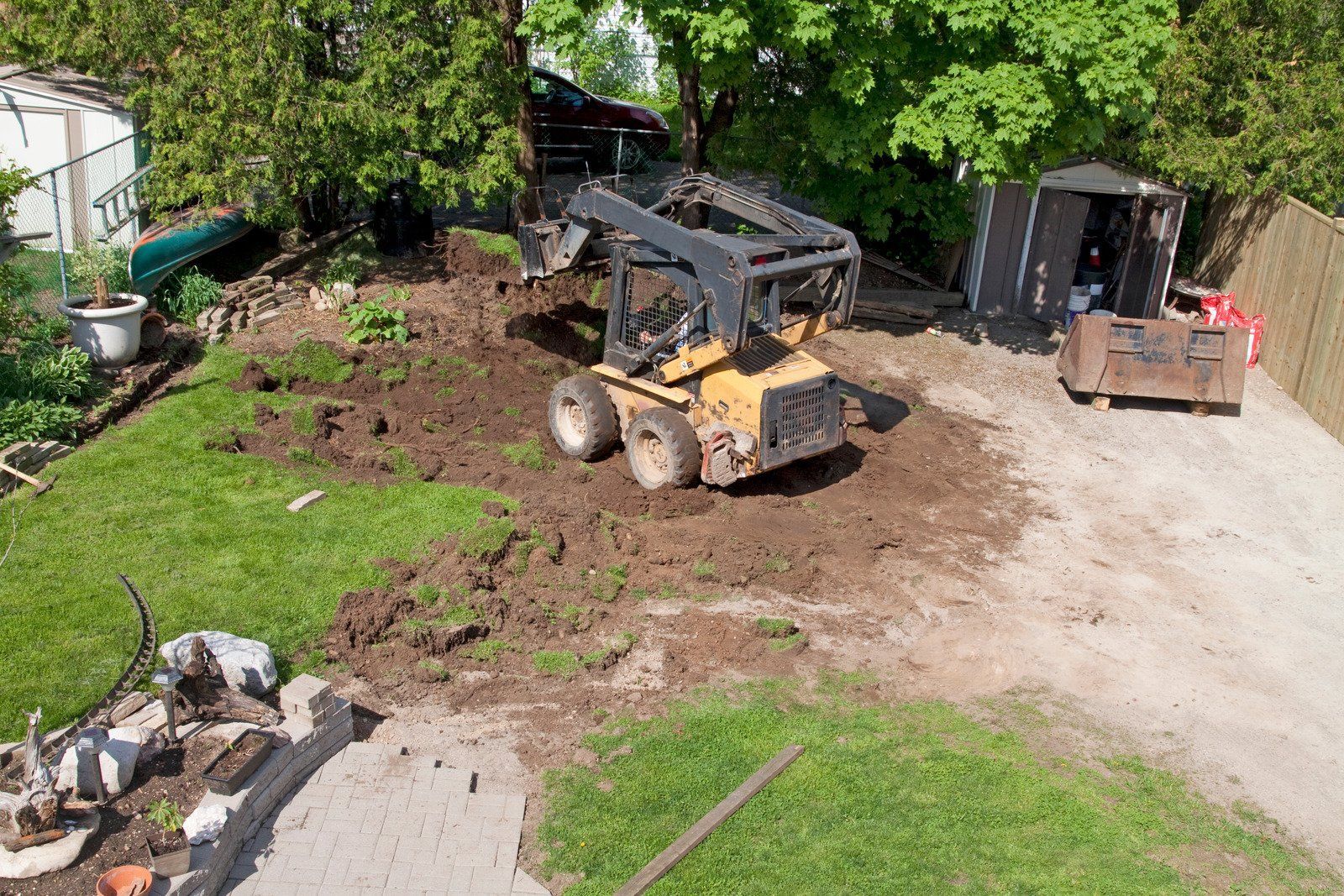 Yellow skid steer grading a dirt area in a residential backyard.