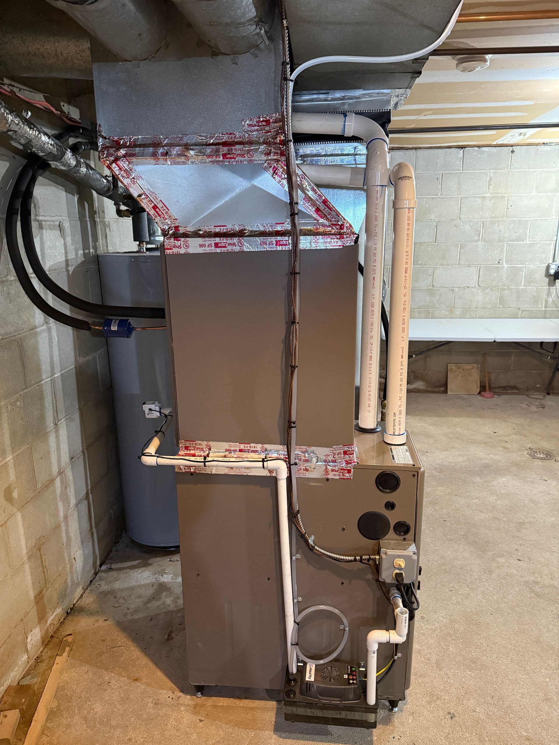 Furnace and water heater in a basement. Metal and gray appliances with white PVC pipes.
