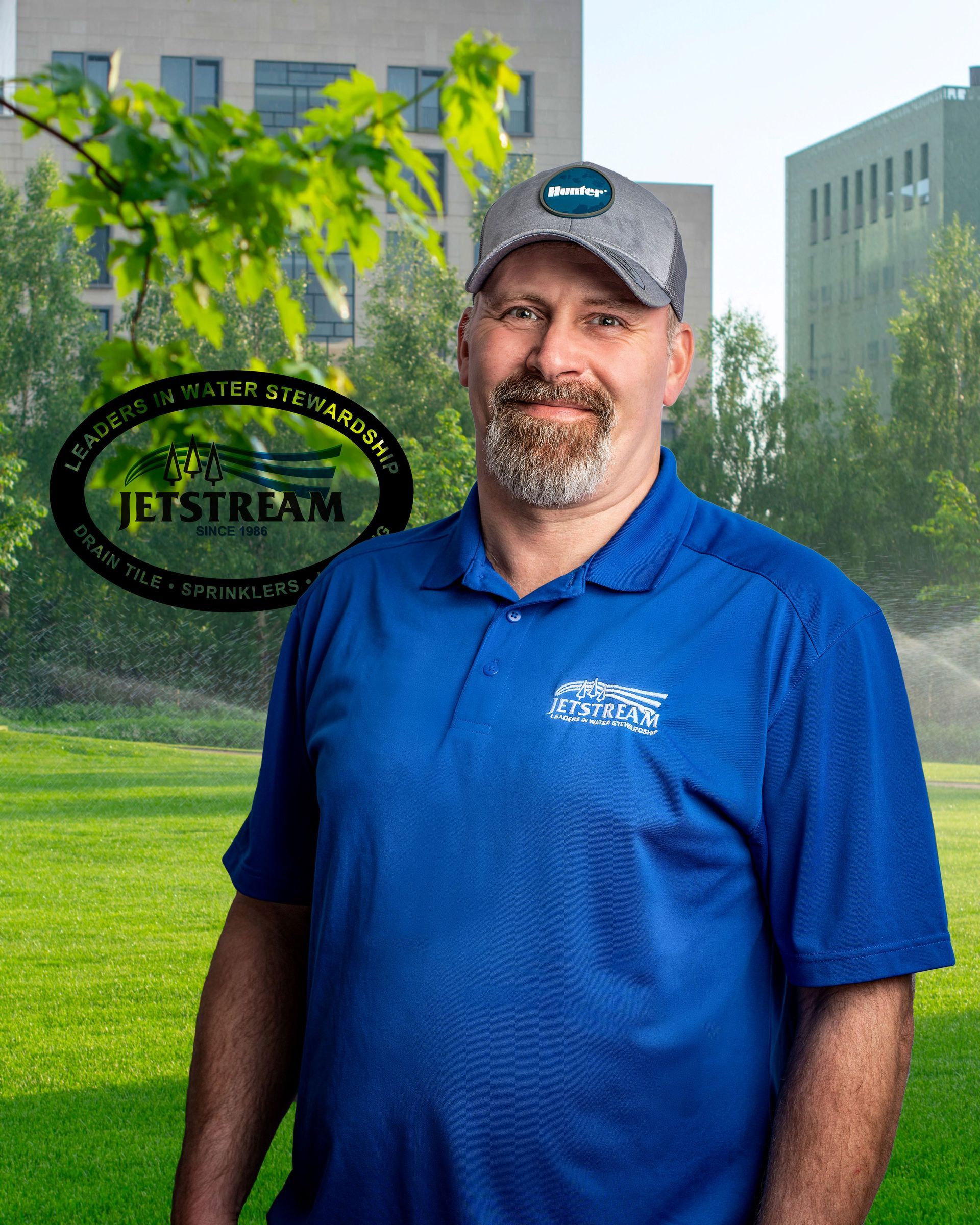 A man in a blue shirt and hat is standing in front of a sprinkler.