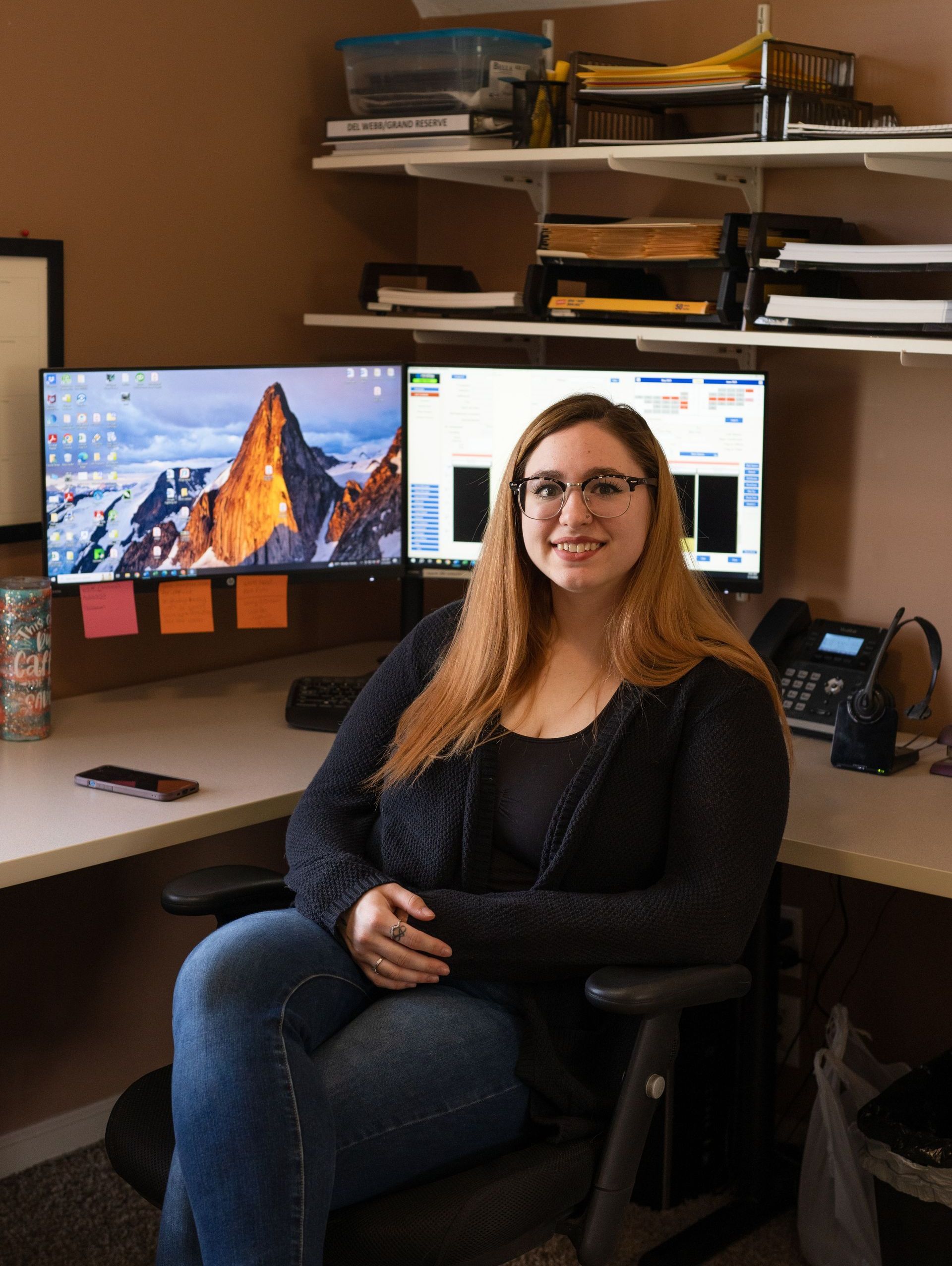 A woman is sitting in an office chair in front of two computer monitors.