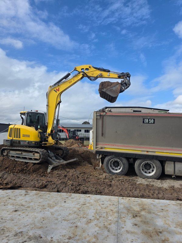 Yellow excavator dumping dirt into a dump truck at a muddy construction site under a blue sky