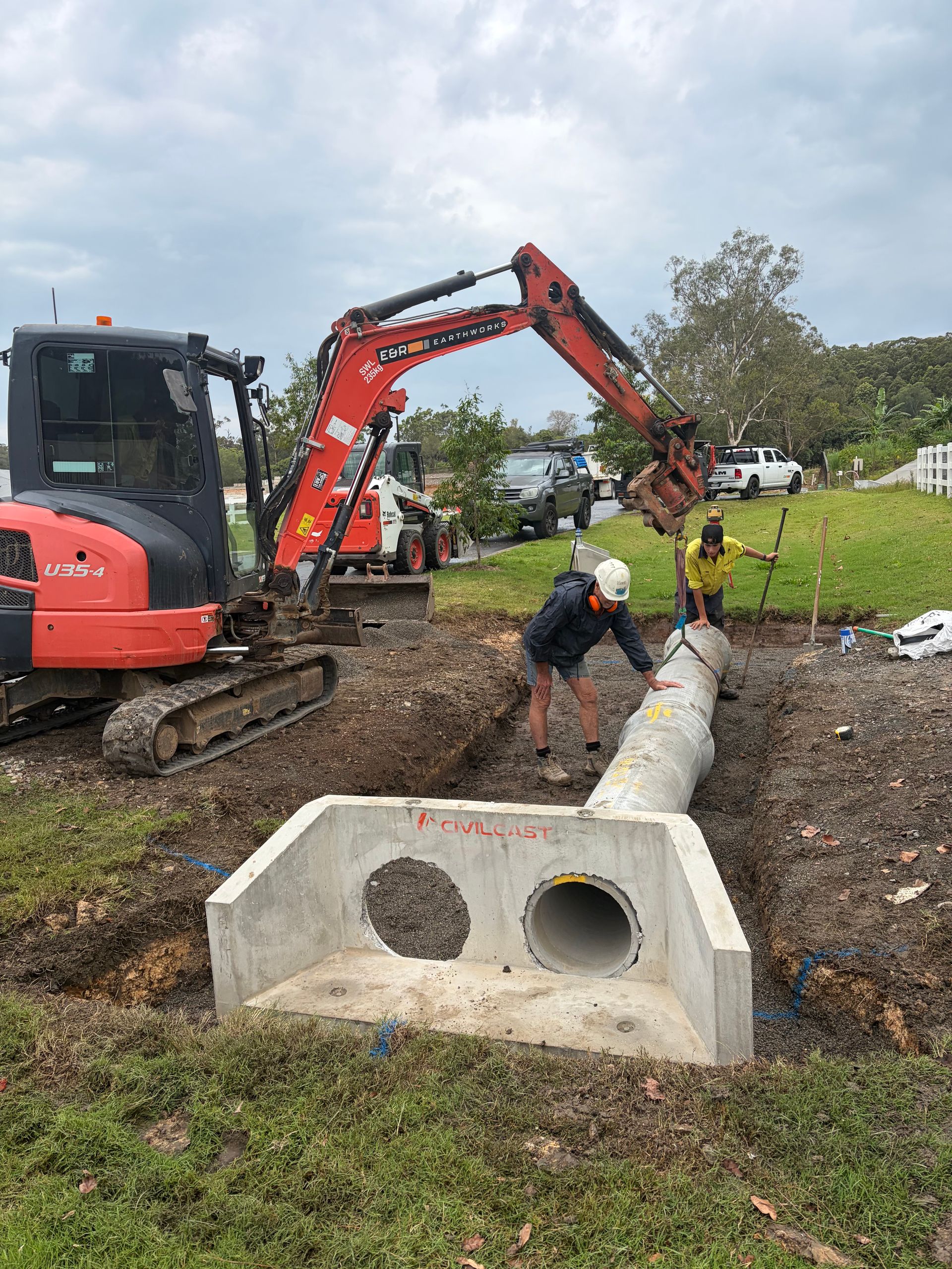Construction crew installing a concrete pipe with an excavator at a muddy work site.