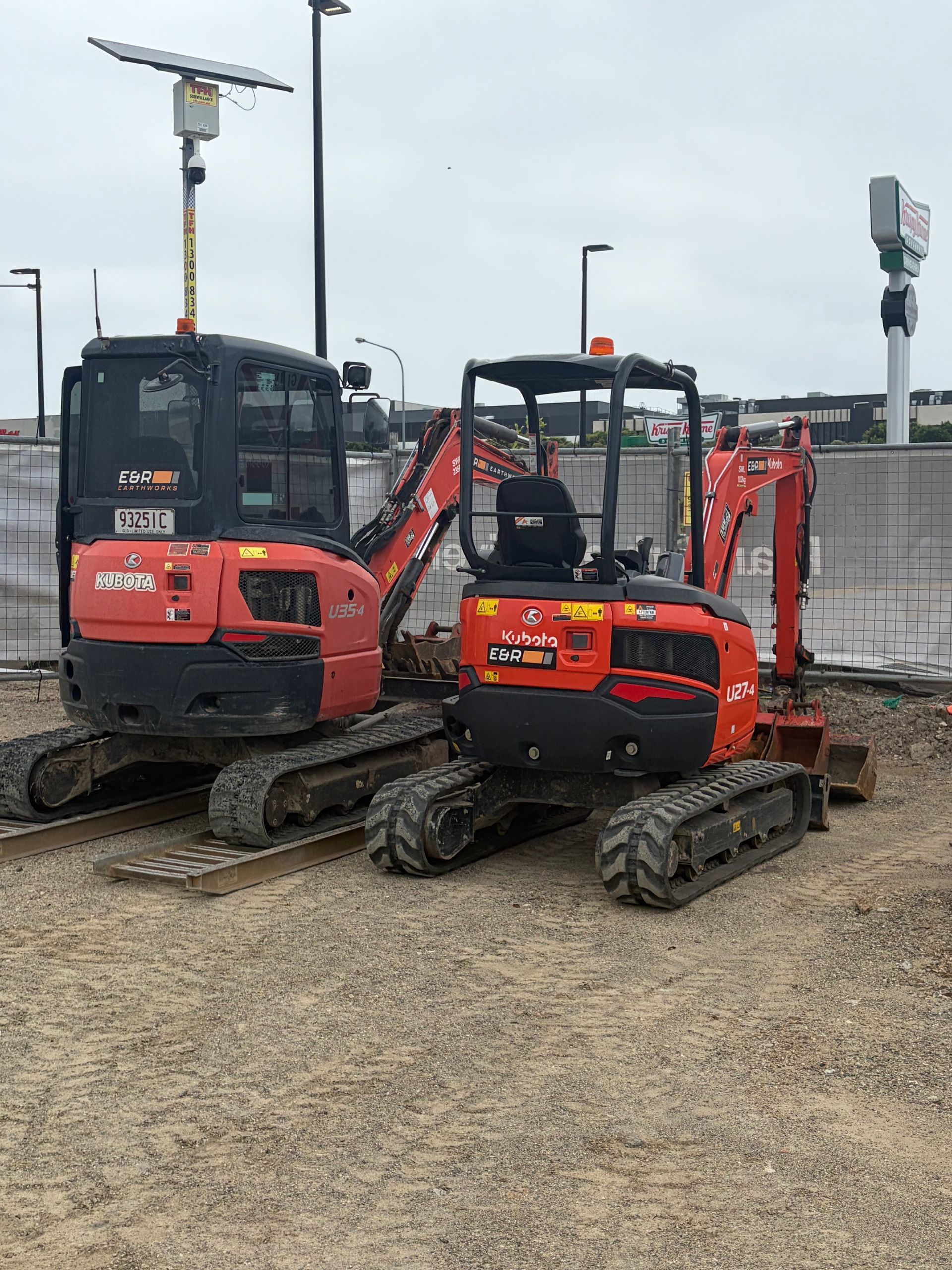 Two red mini excavators parked on a gravel lot under cloudy skies