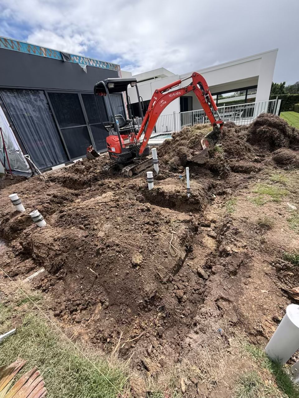 Red excavator digging a large muddy area beside a modern building and black fence