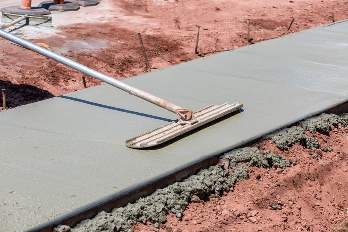 A worker uses a long-handled tool to smooth fresh concrete in a sidewalk form.