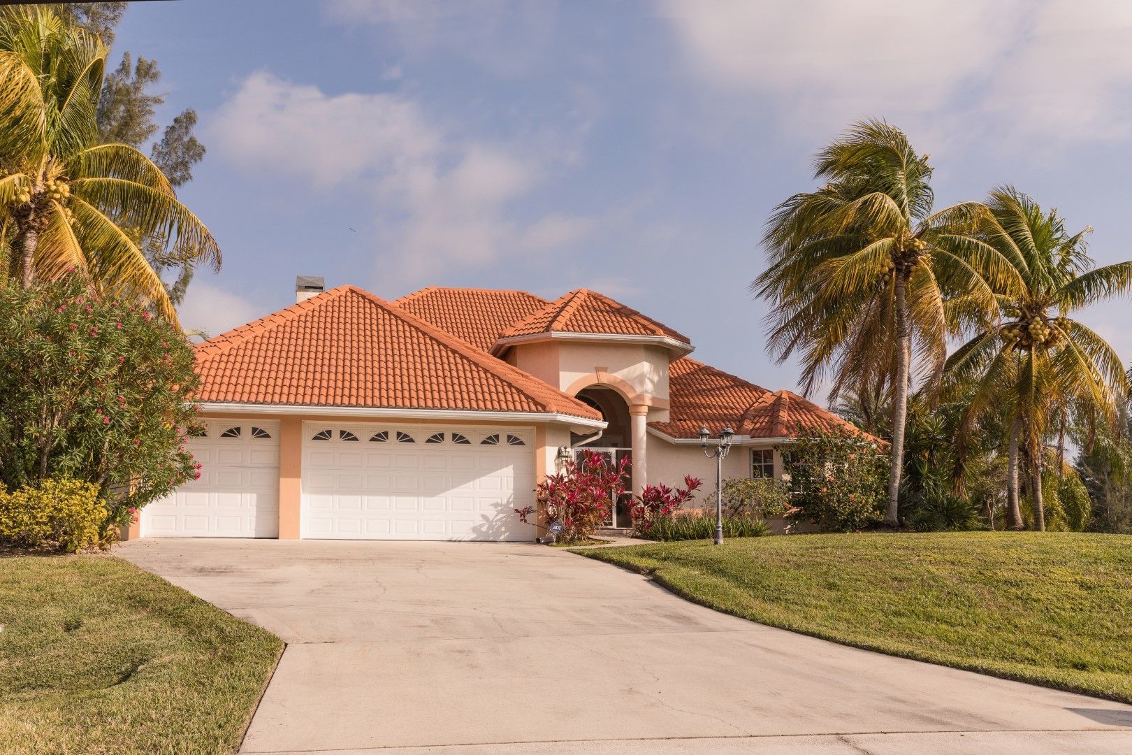 A stucco house with a red tile roof, white garage doors, and a driveway, surrounded by palm trees and green grass.