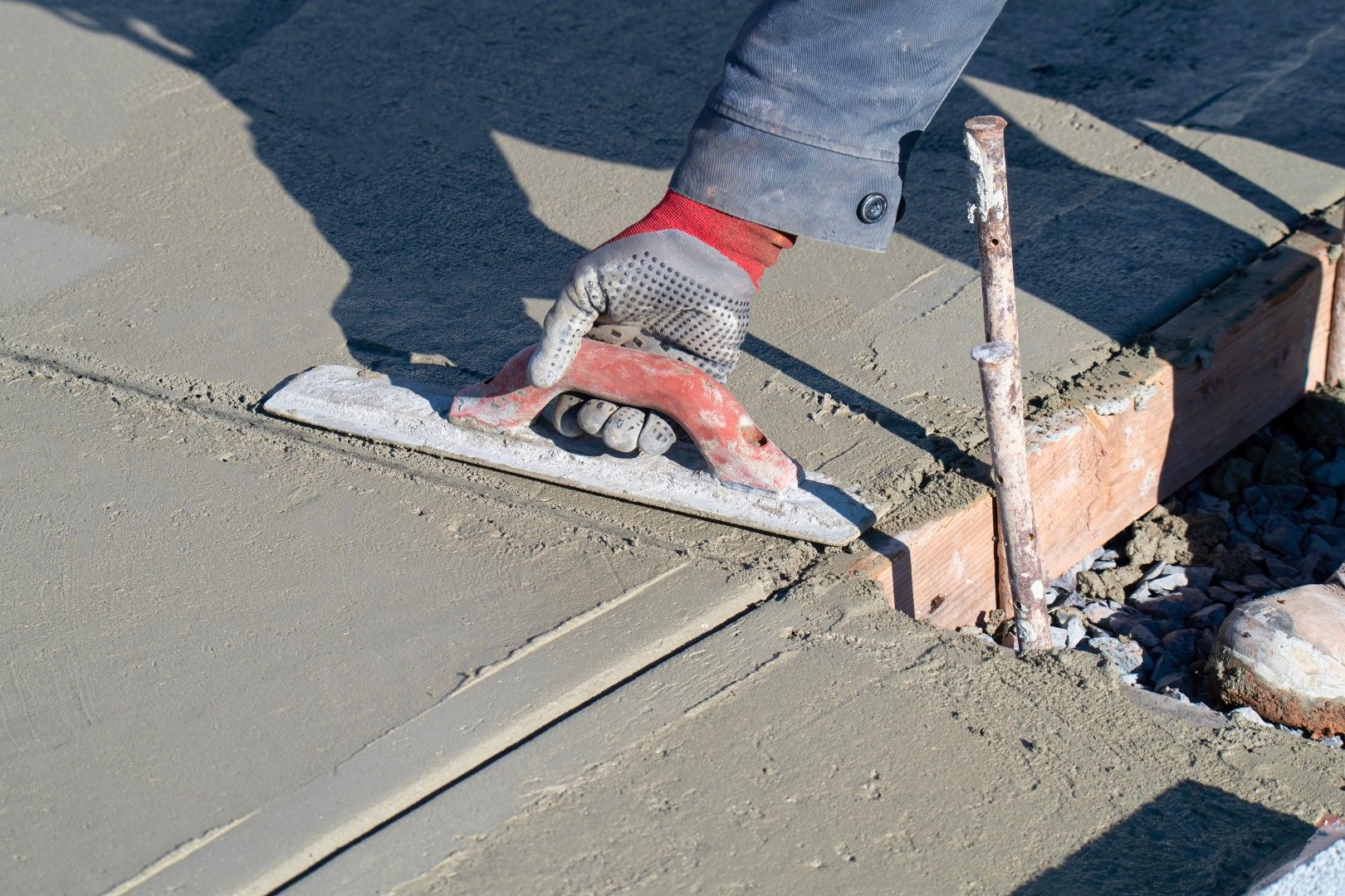 A gloved hand uses a trowel to smooth wet concrete along a brick border outdoors.