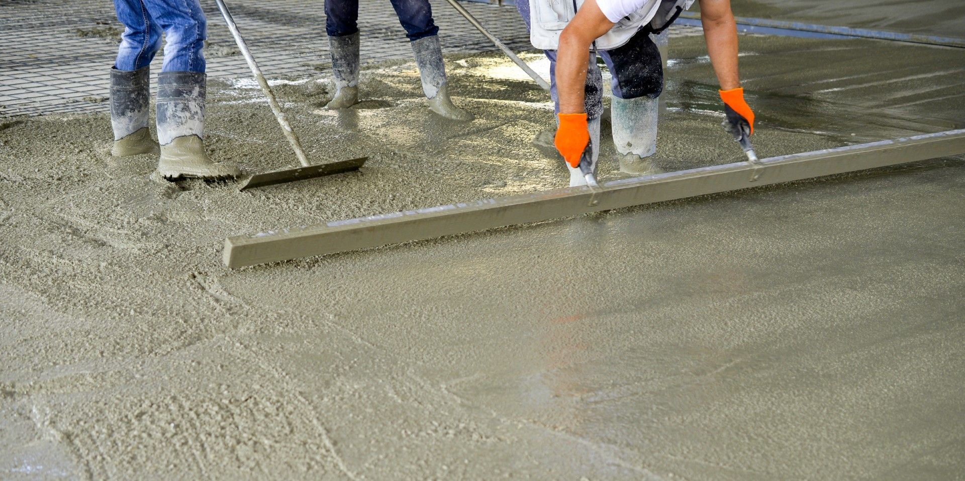 Workers smoothing wet concrete with tools, likely at a construction site. They are wearing boots and gloves.
