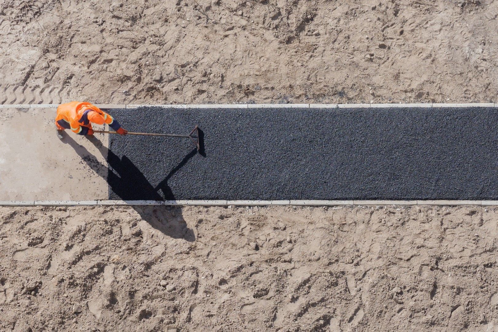 Worker in orange vest rolling black asphalt strip on sandy ground. Concrete border visible.