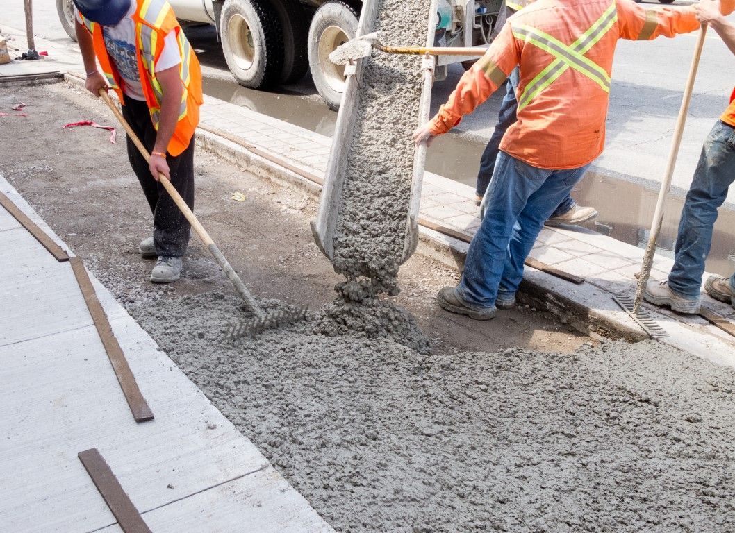 Construction workers pouring wet concrete from a truck, leveling it with rakes, orange safety vests, city sidewalk.