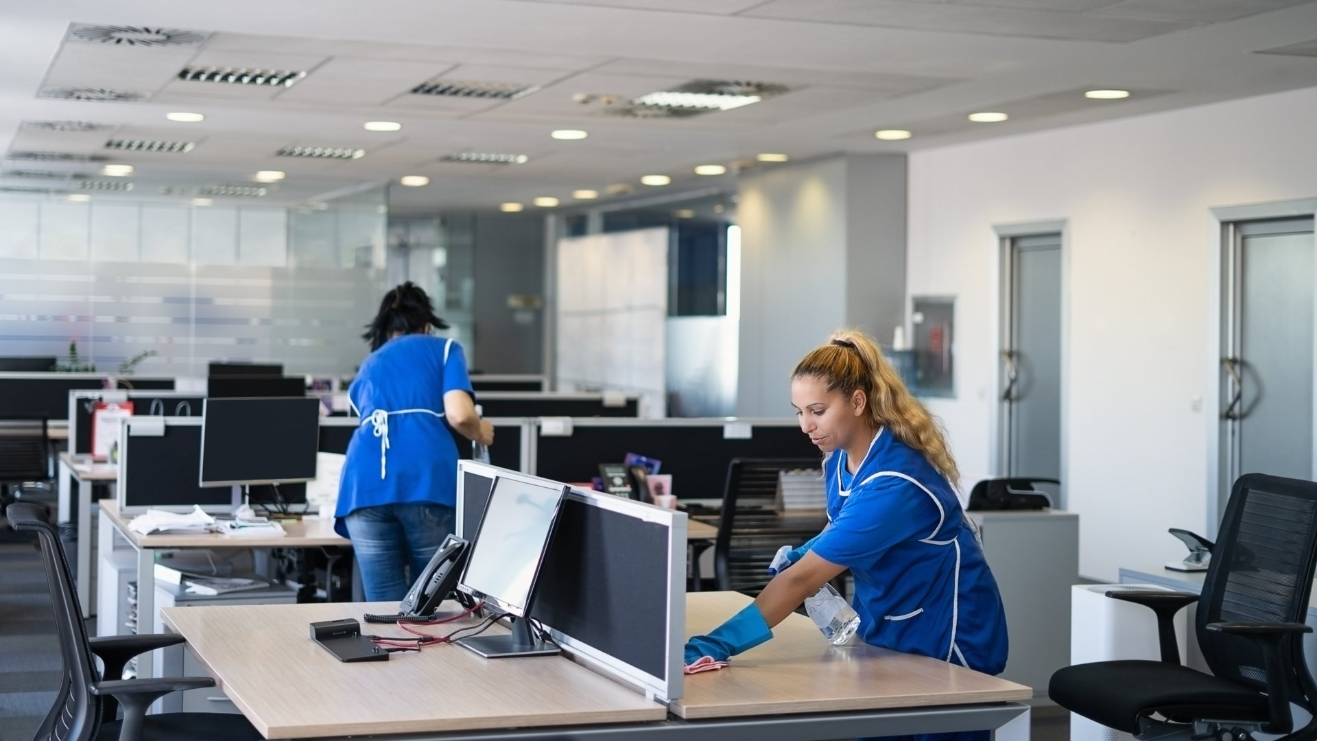 Two women are cleaning desks in an office.