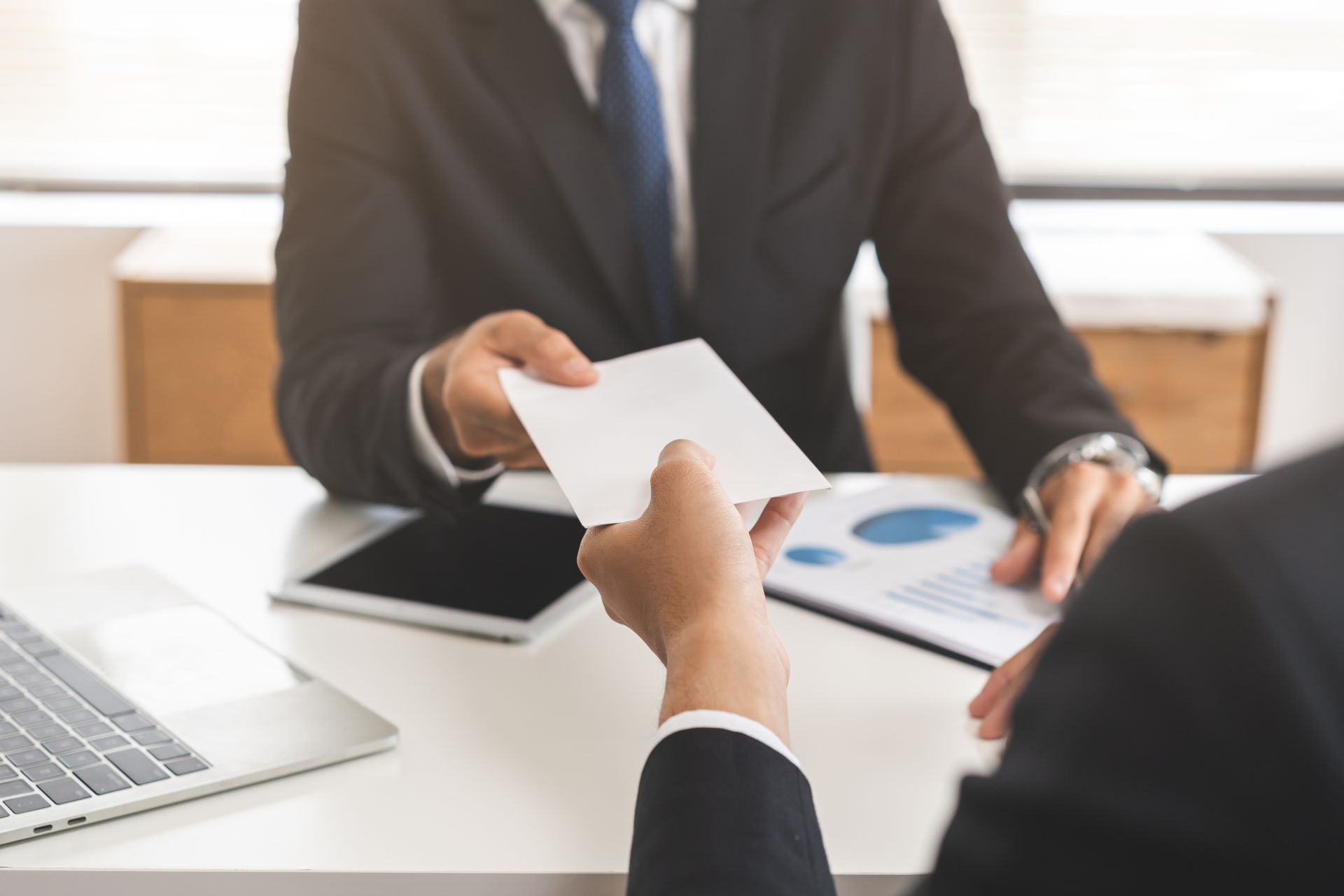 Business meeting handoff of a document across a desk with laptop and charts nearby