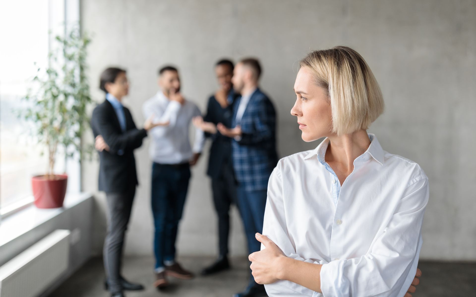 Woman in white shirt standing in foreground, arms crossed, with coworkers talking blurred in the background.
