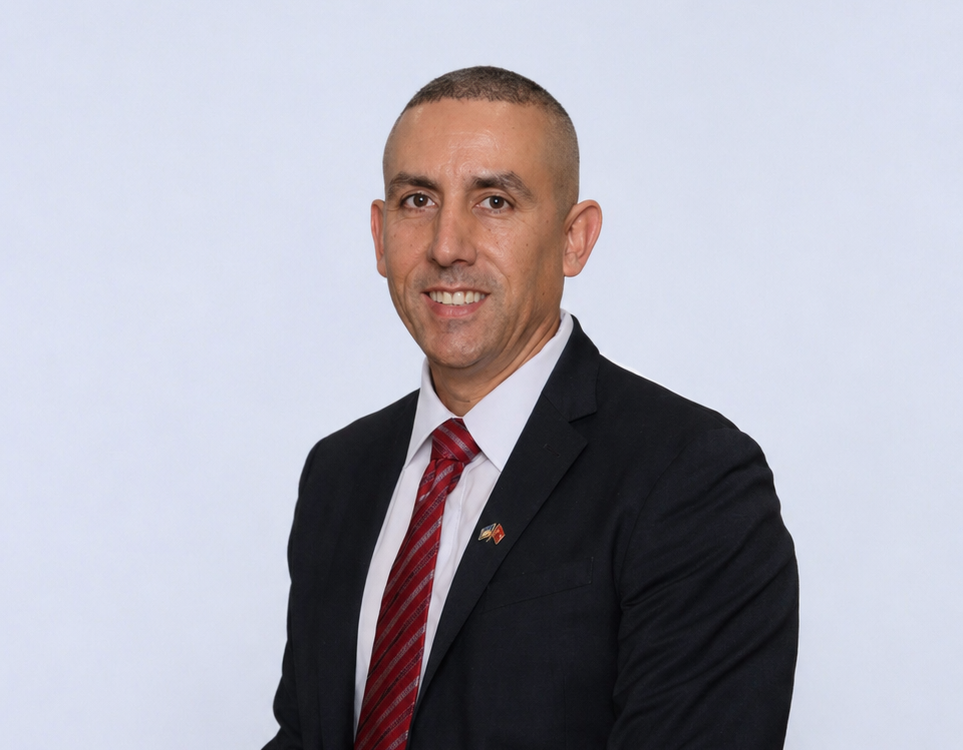Man in a dark suit with a red striped tie, standing indoors and smiling toward the camera