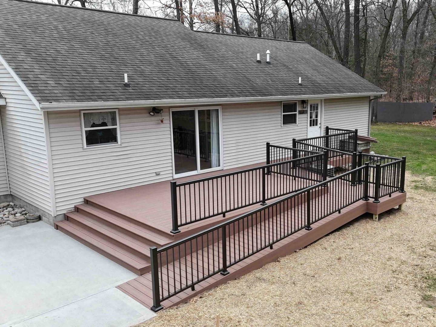 House with a composite deck, ramp, and dark railings. Brown steps and a concrete patio lead to the deck.