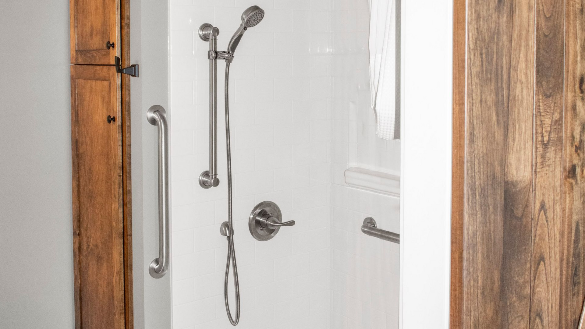 A walk-in shower with white walls, a handheld showerhead on a slider bar, a water control handle, and silver grab bars.