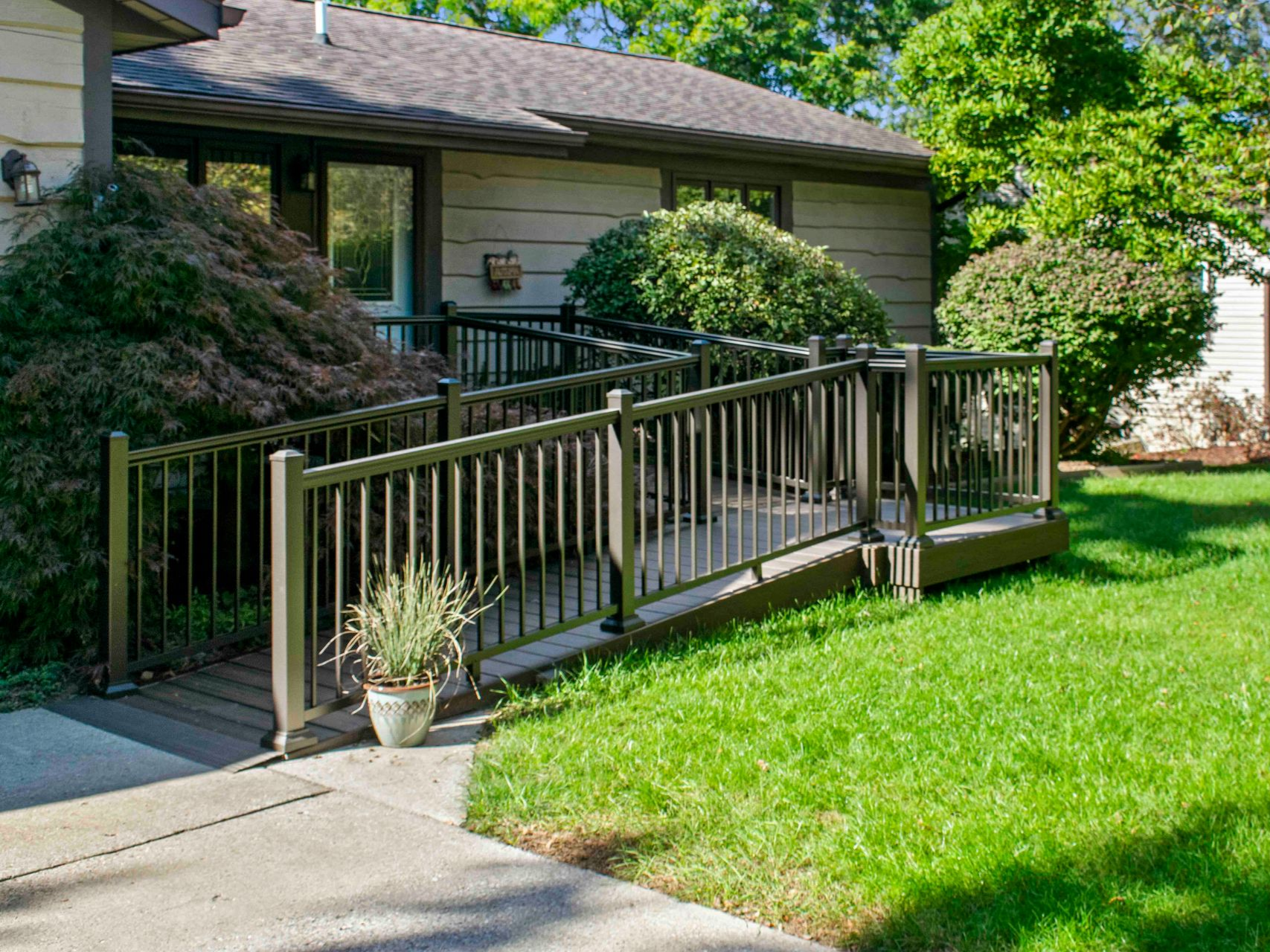 Ramped entrance to a house with dark railings and a small porch, surrounded by greenery and lawn.