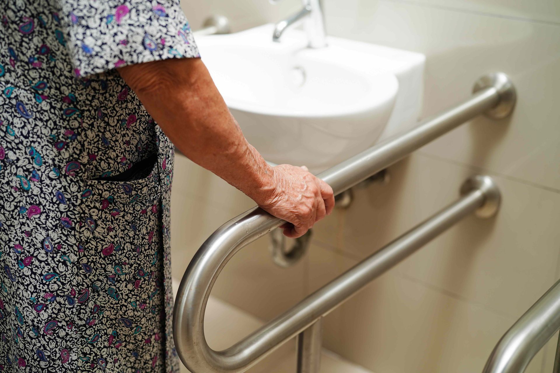 Person grasping a stainless steel handrail next to a sink in a bathroom.