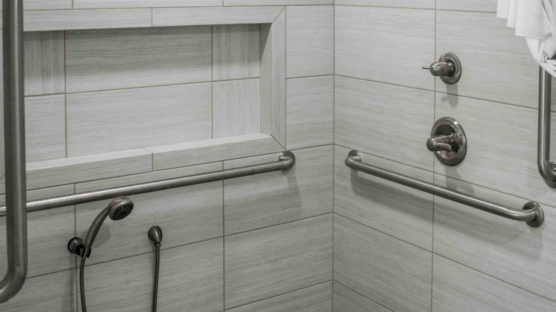 An accessible shower stall featuring light gray tiled walls, stainless steel grab bars, a handheld showerhead, and controls.