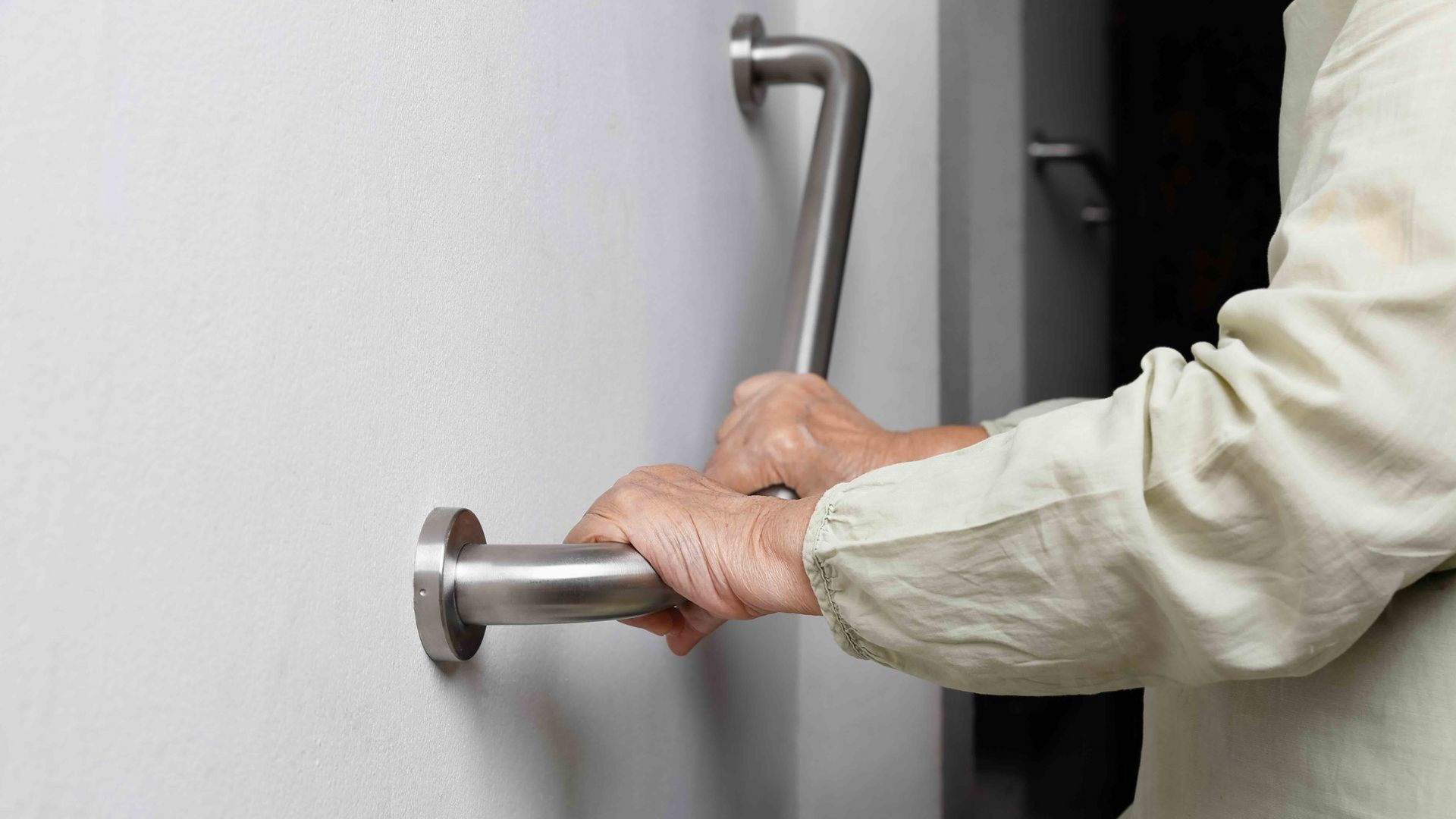 A person's hands gripping a stainless steel grab bar mounted on a white wall for support.