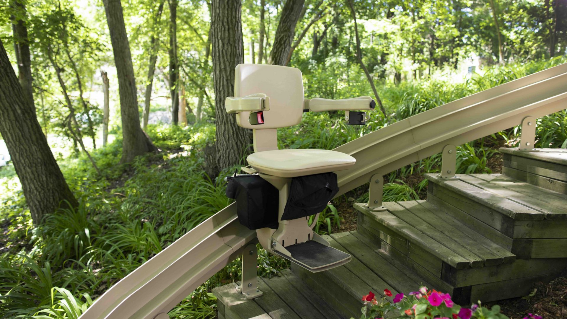 An outdoor stair lift with a tan seat mounted on a metal rail along wooden stairs in a wooded garden setting.