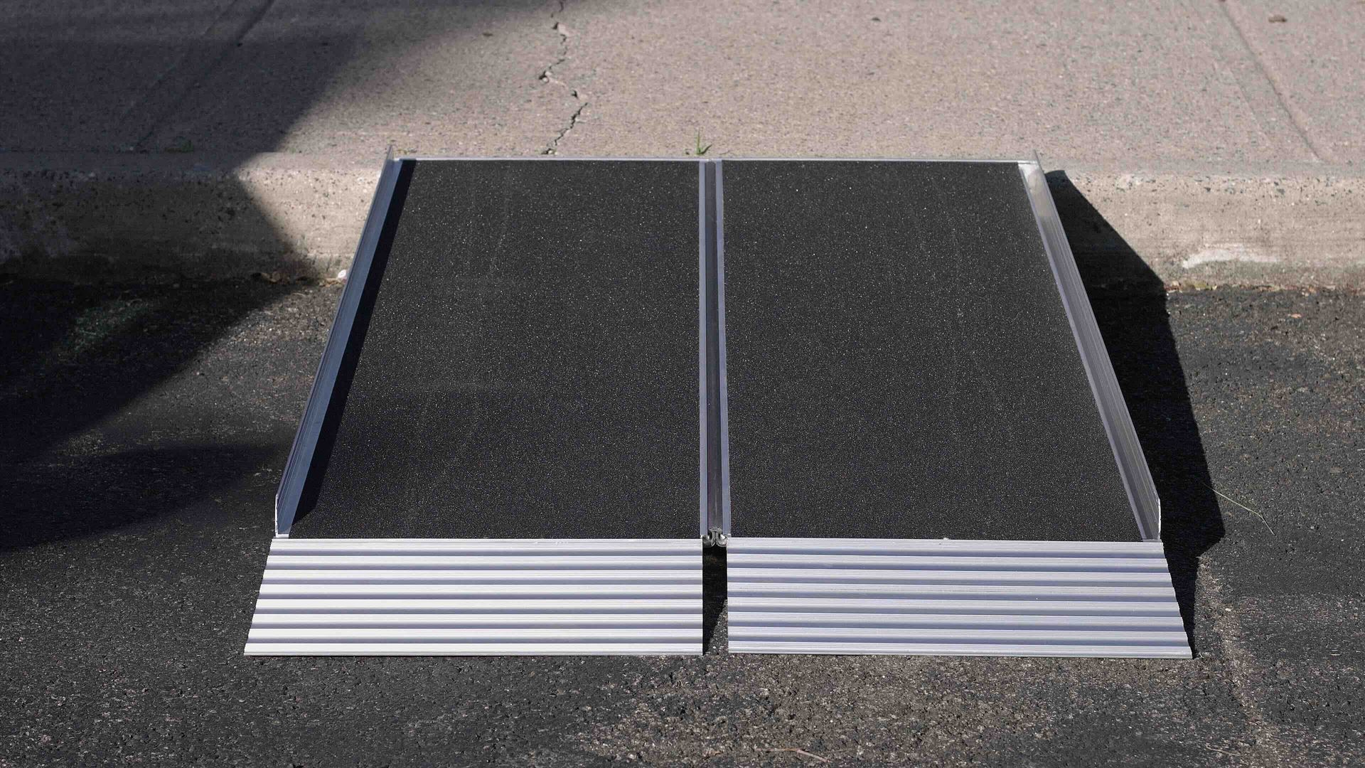 A portable two-piece aluminum wheelchair ramp with a black non-slip surface, resting against a sidewalk curb.