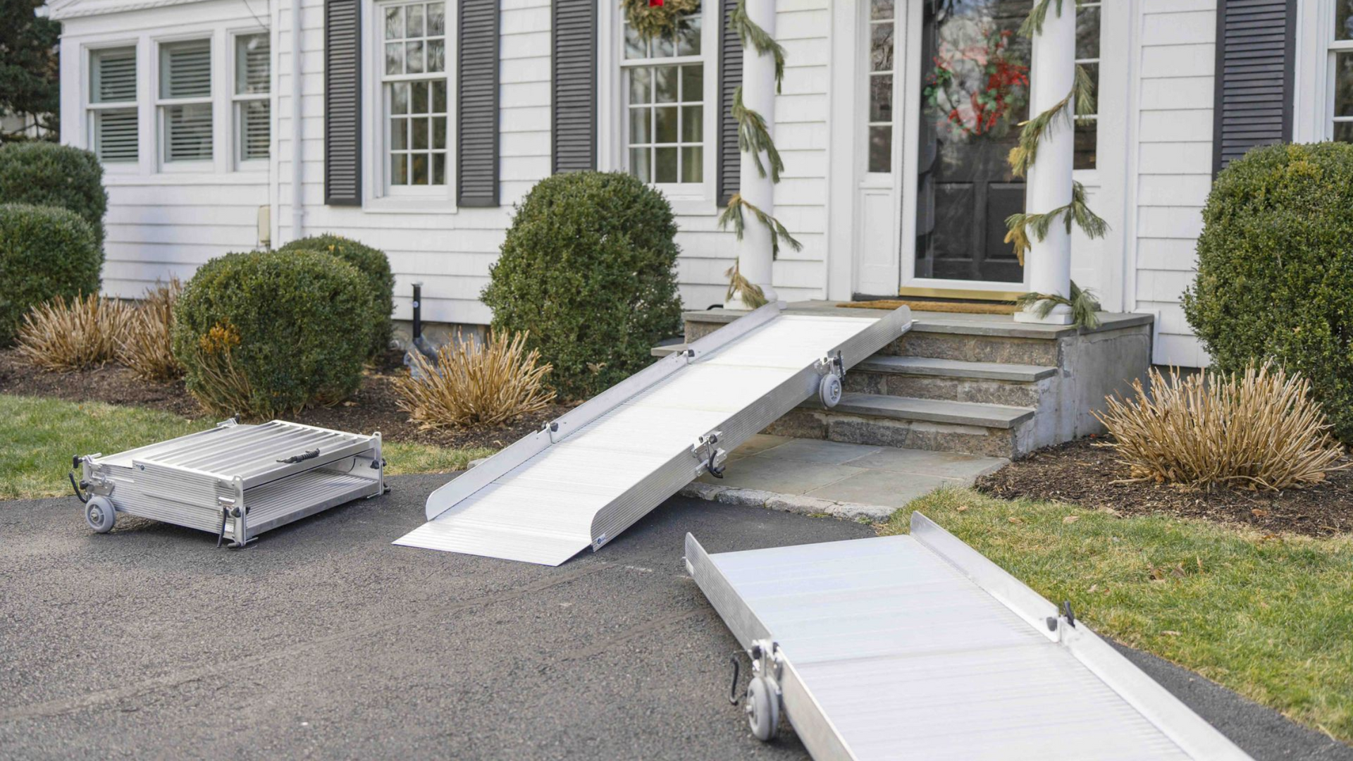 Portable aluminum ramps are displayed outside a home, with one ramp installed on the front steps and two others resting nearby.