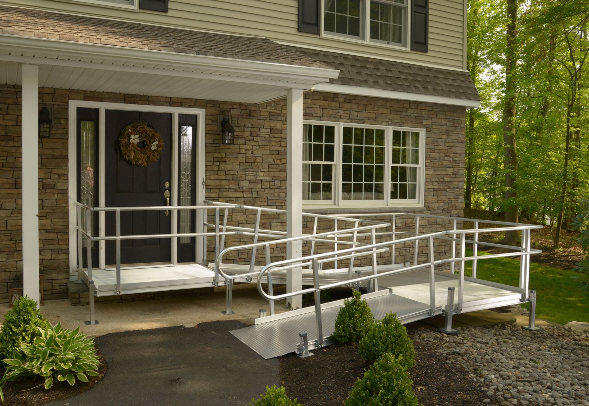 Exterior of a house with an aluminum ramp for accessibility. Ramp leads to the front door; stone exterior and green bushes.