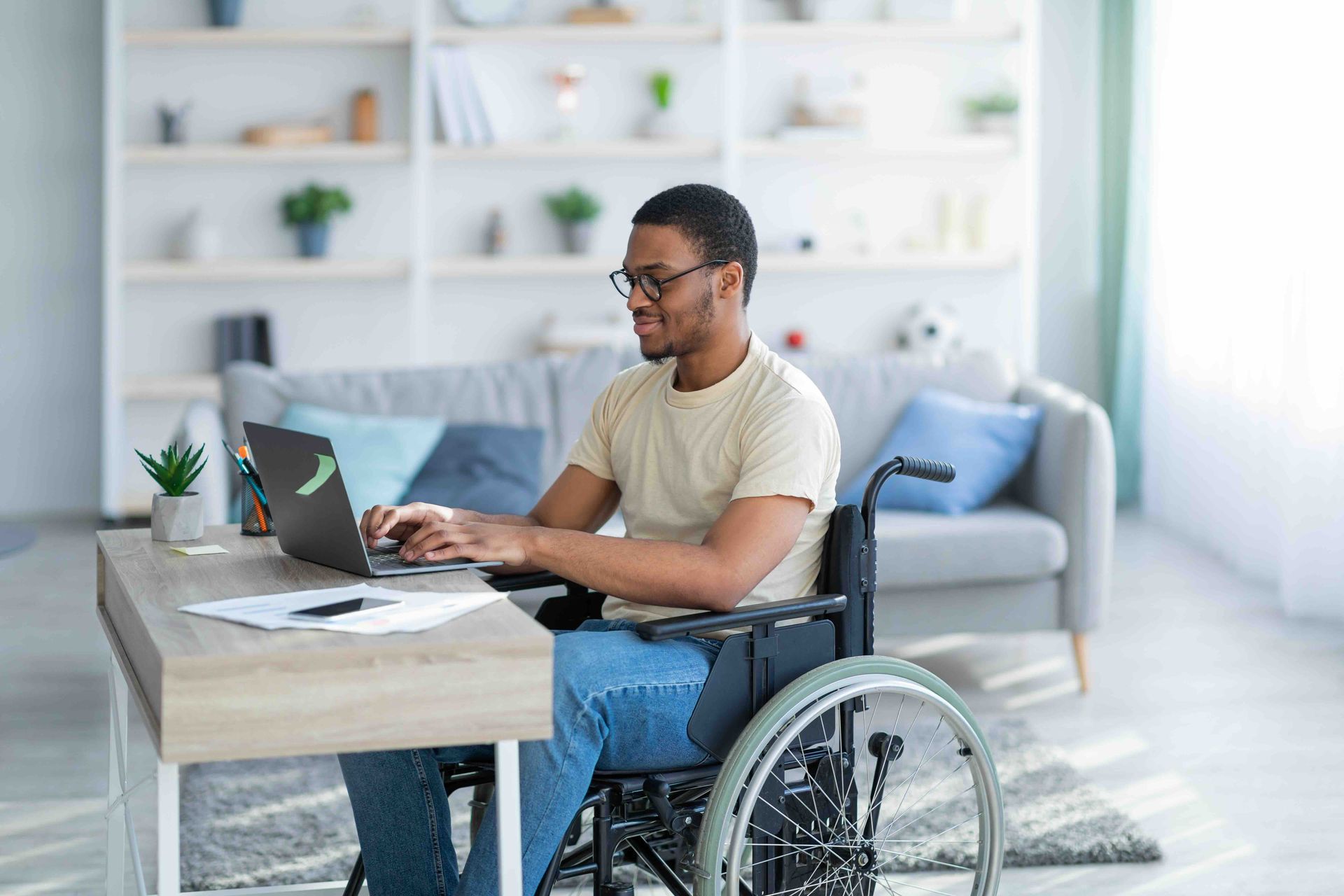 Man in a wheelchair working on a laptop at a desk in a living room, smiling.