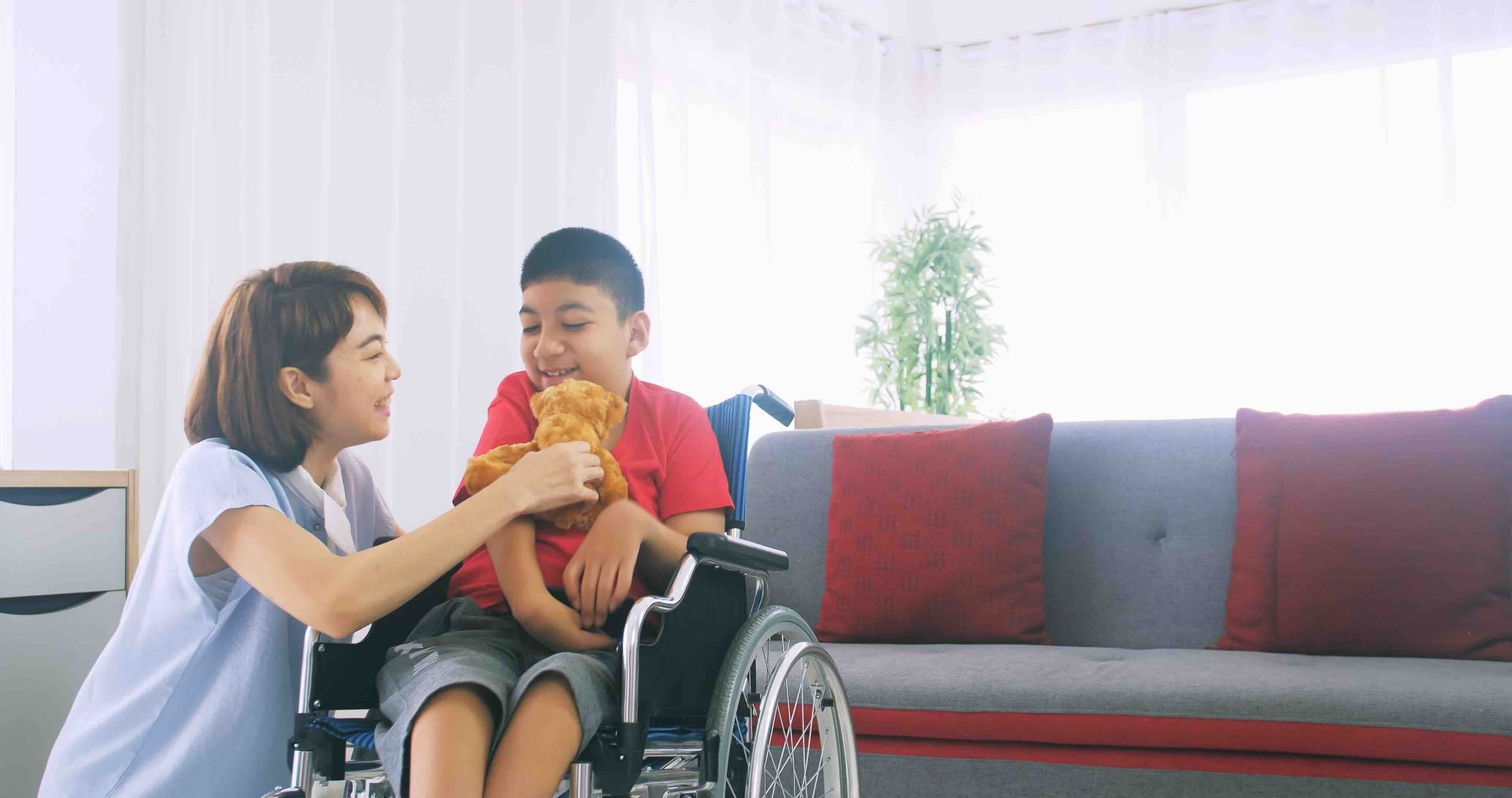 A woman is handing a teddy bear to a boy in a wheelchair.