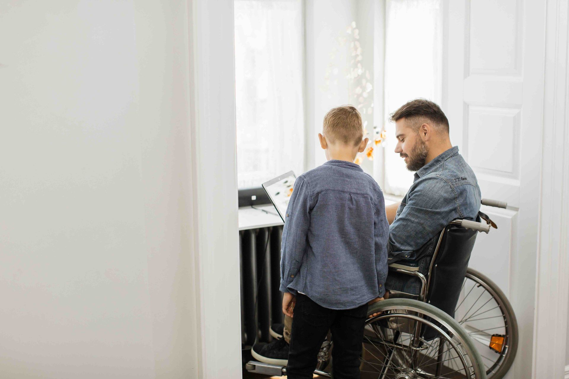 A man in a wheelchair is looking at a laptop with a boy standing next to him.