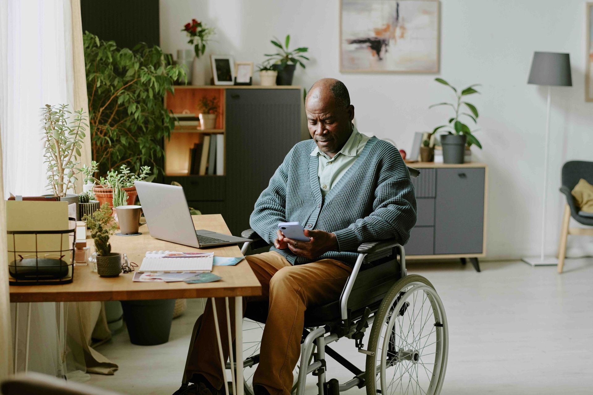 Older Black man in wheelchair using smartphone at desk with laptop and plants in home.