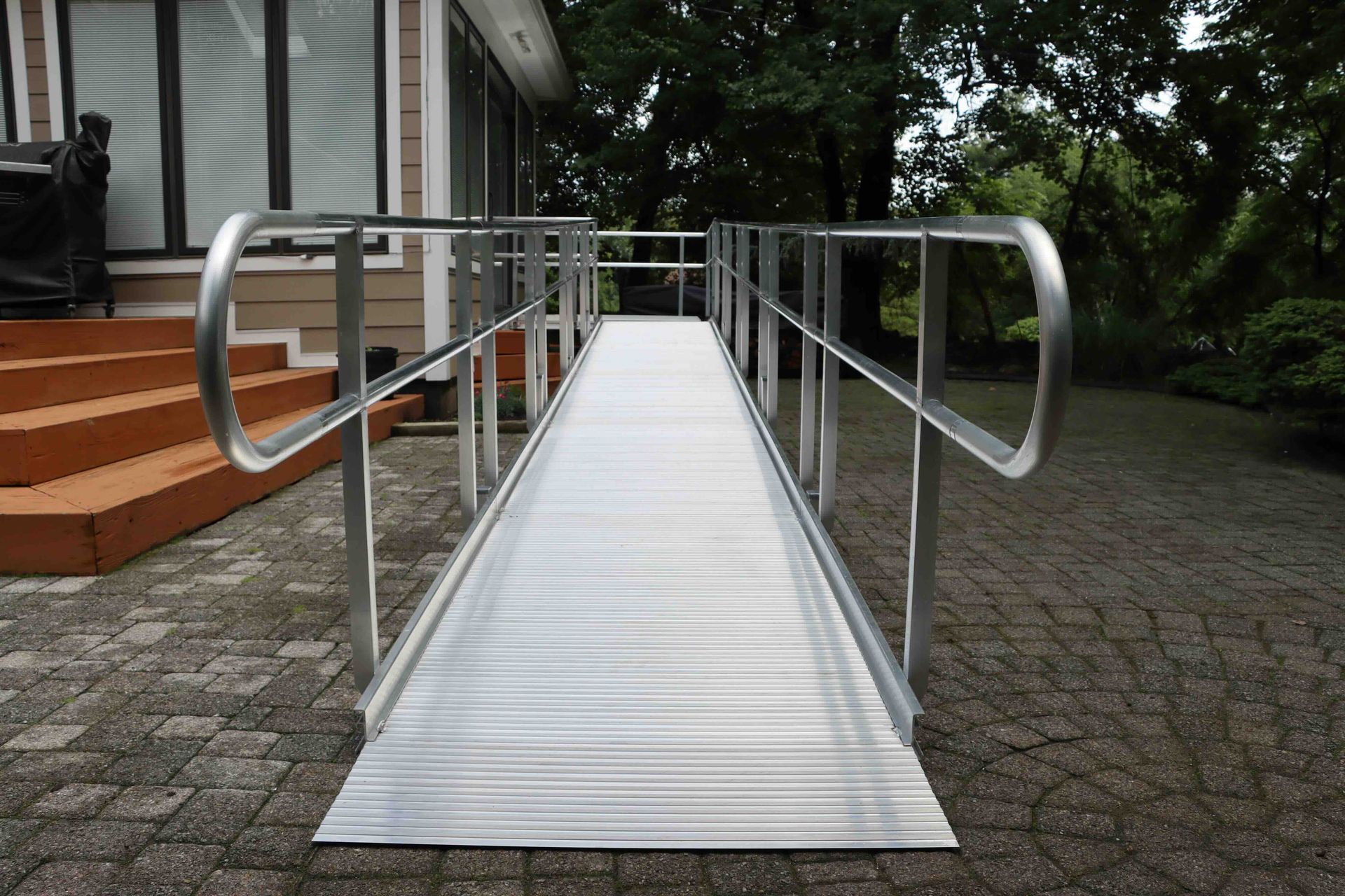 Aluminum ramp with handrails leading to a building's entrance. Outdoor setting with greenery.