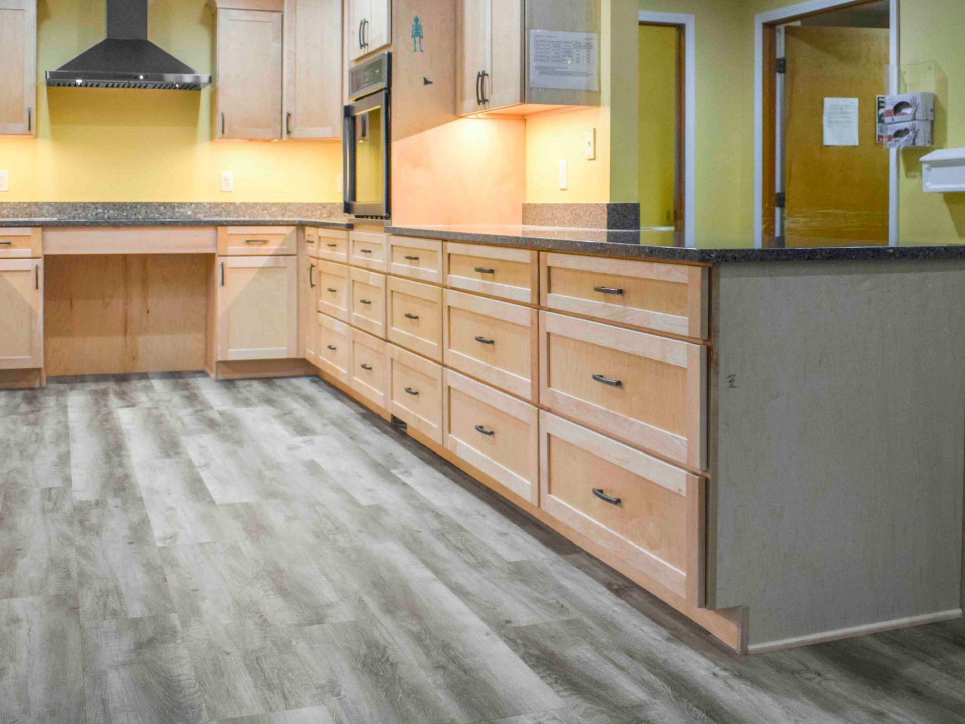 Kitchen with wood cabinets, black countertops, and light gray flooring. Yellow walls, and a black range hood.