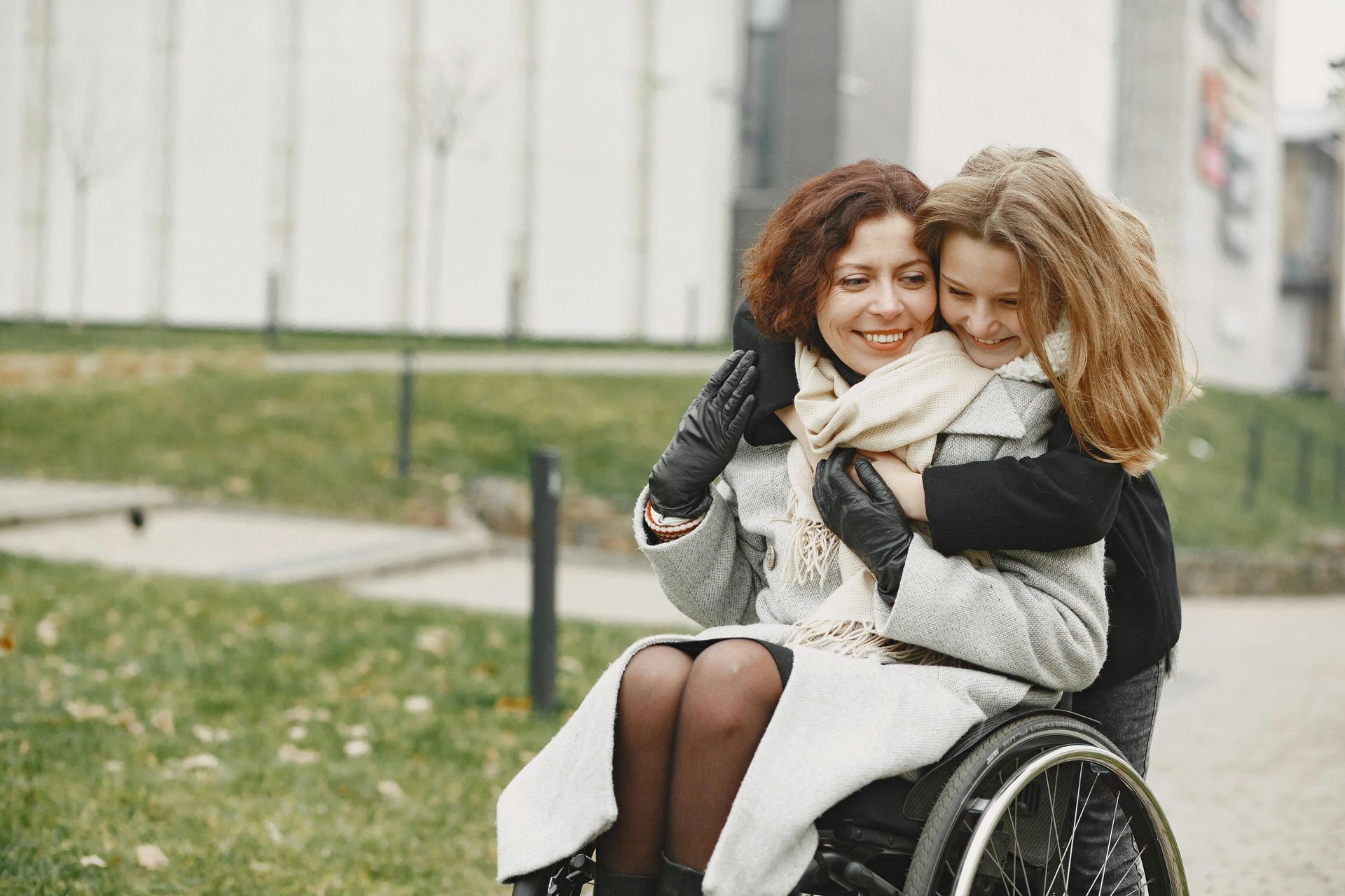 Woman in wheelchair embraces a child outdoors, both smiling, wearing coats and scarves.