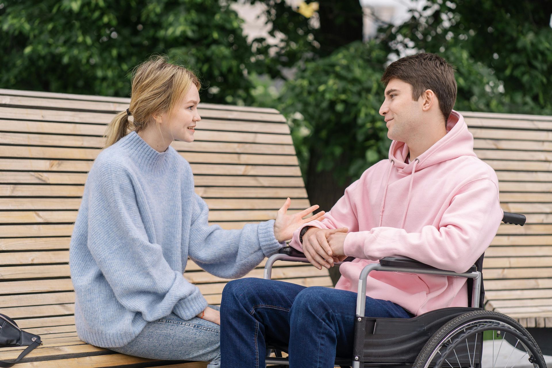 Woman and man in wheelchair talking on a park bench. Man in pink hoodie, woman in blue sweater; both smiling.