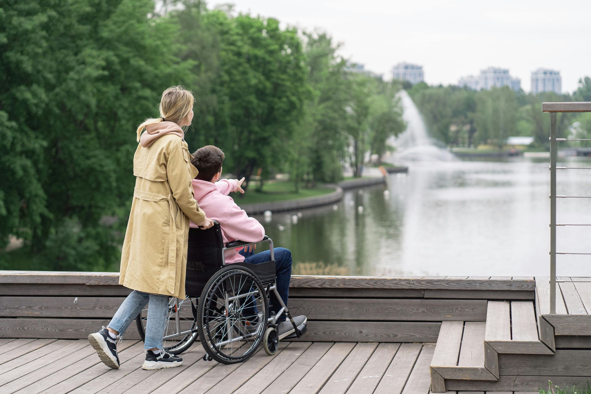Woman in yellow coat assists person in wheelchair pointing at a park fountain by a lake.