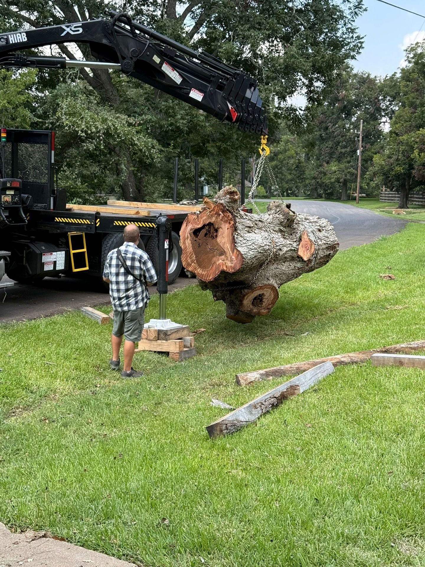 A crane mounted on a flatbed truck lifts a large, hollowed-out tree log in a residential grassy area.