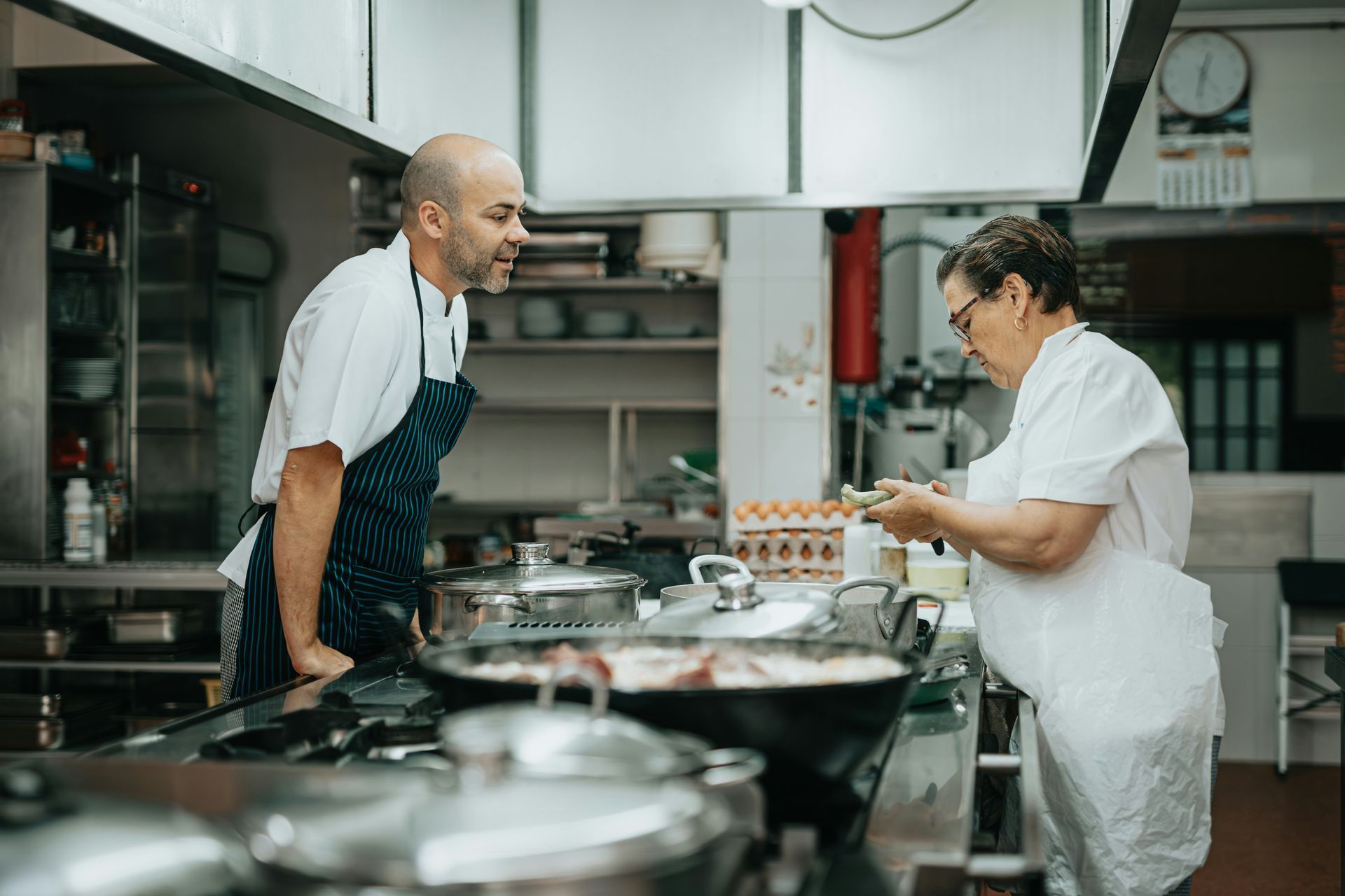 Two chefs in a commercial kitchen; one looking on, one working over a pan, light interior.
