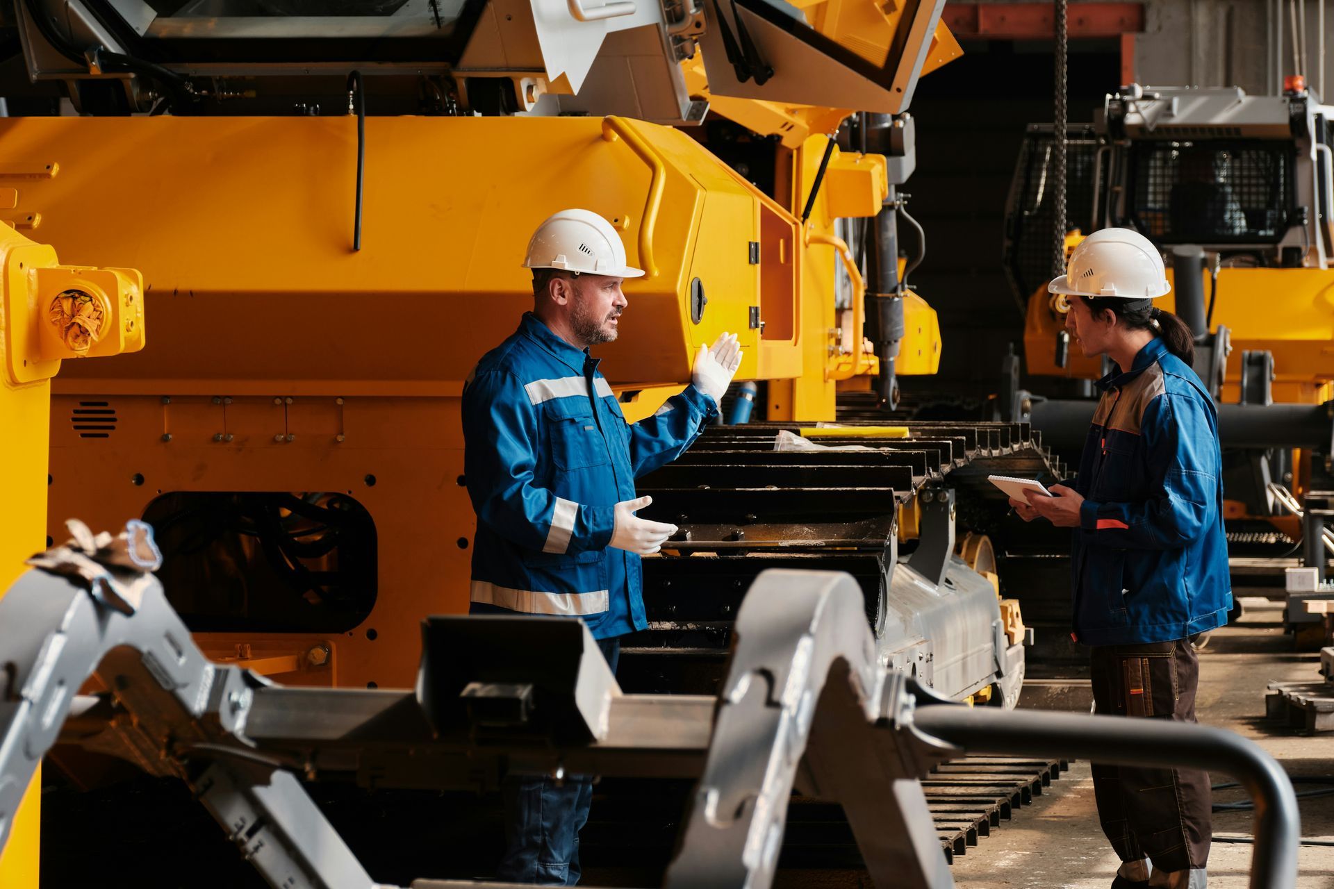 Two workers in blue uniforms and white helmets discussing in a yellow machinery factory.
