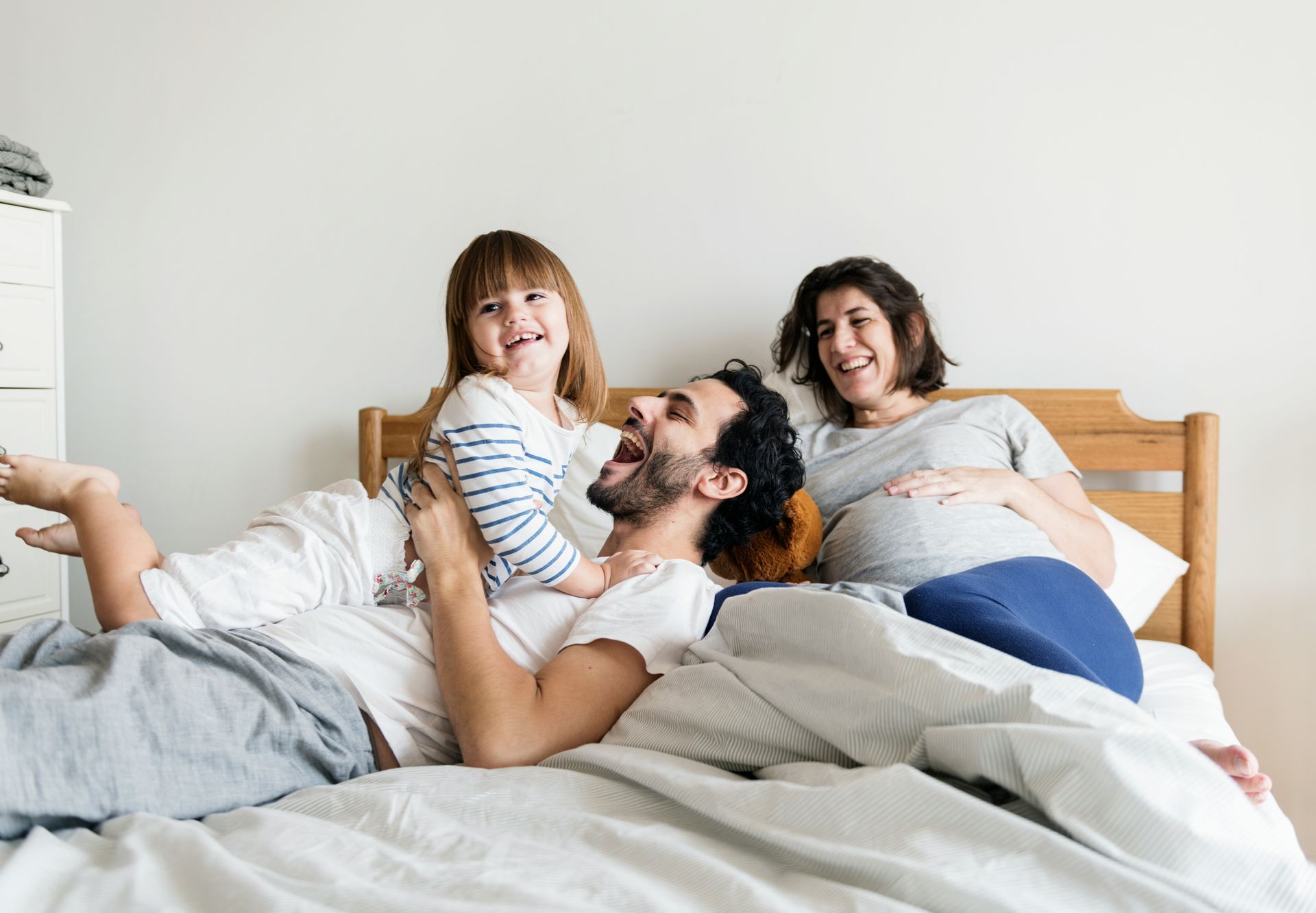 Family of three on a bed: Man laughing, holding a child, pregnant woman smiling, hand on belly.