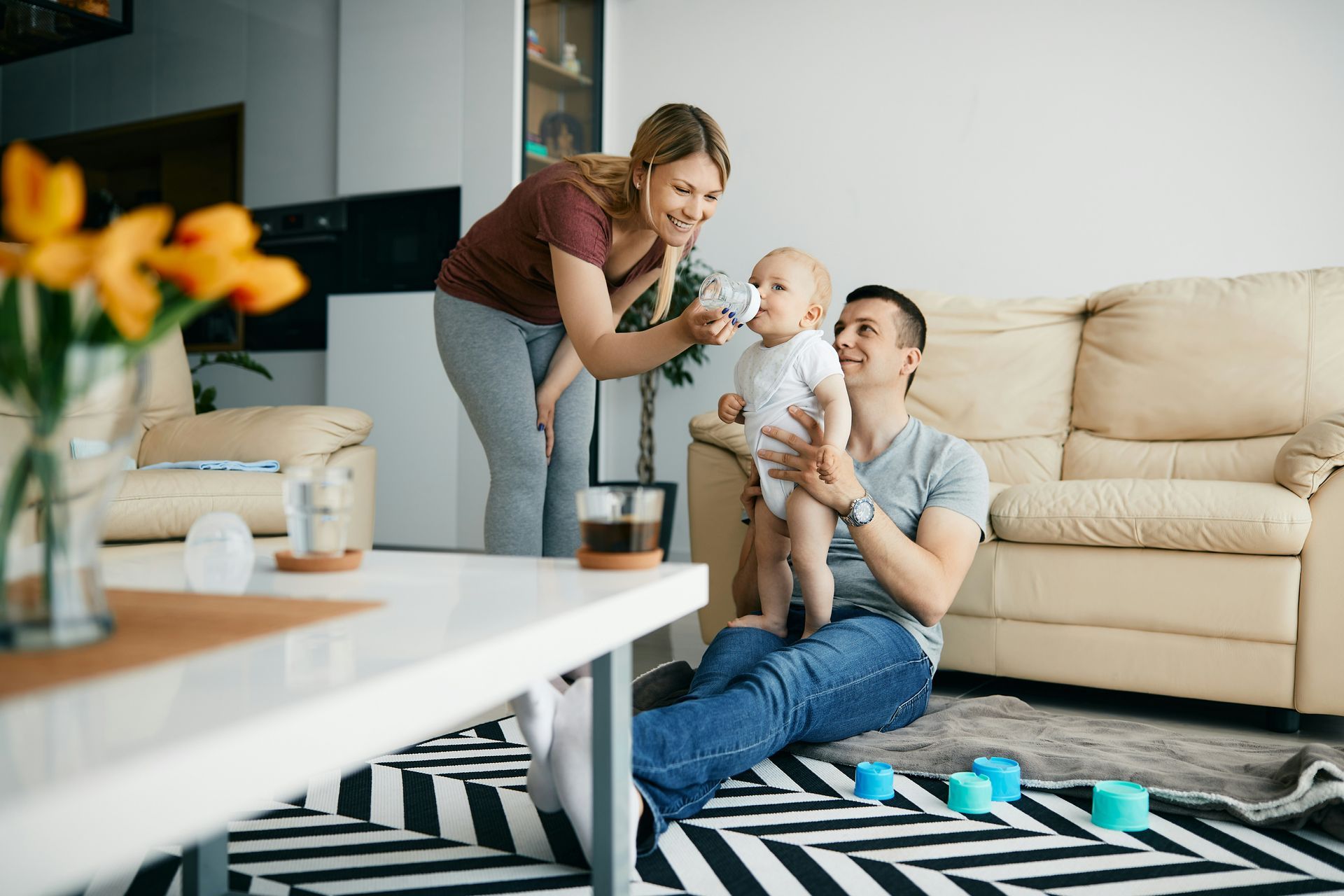 Woman feeding baby a bottle as man watches. Family in modern living room, with yellow flowers.