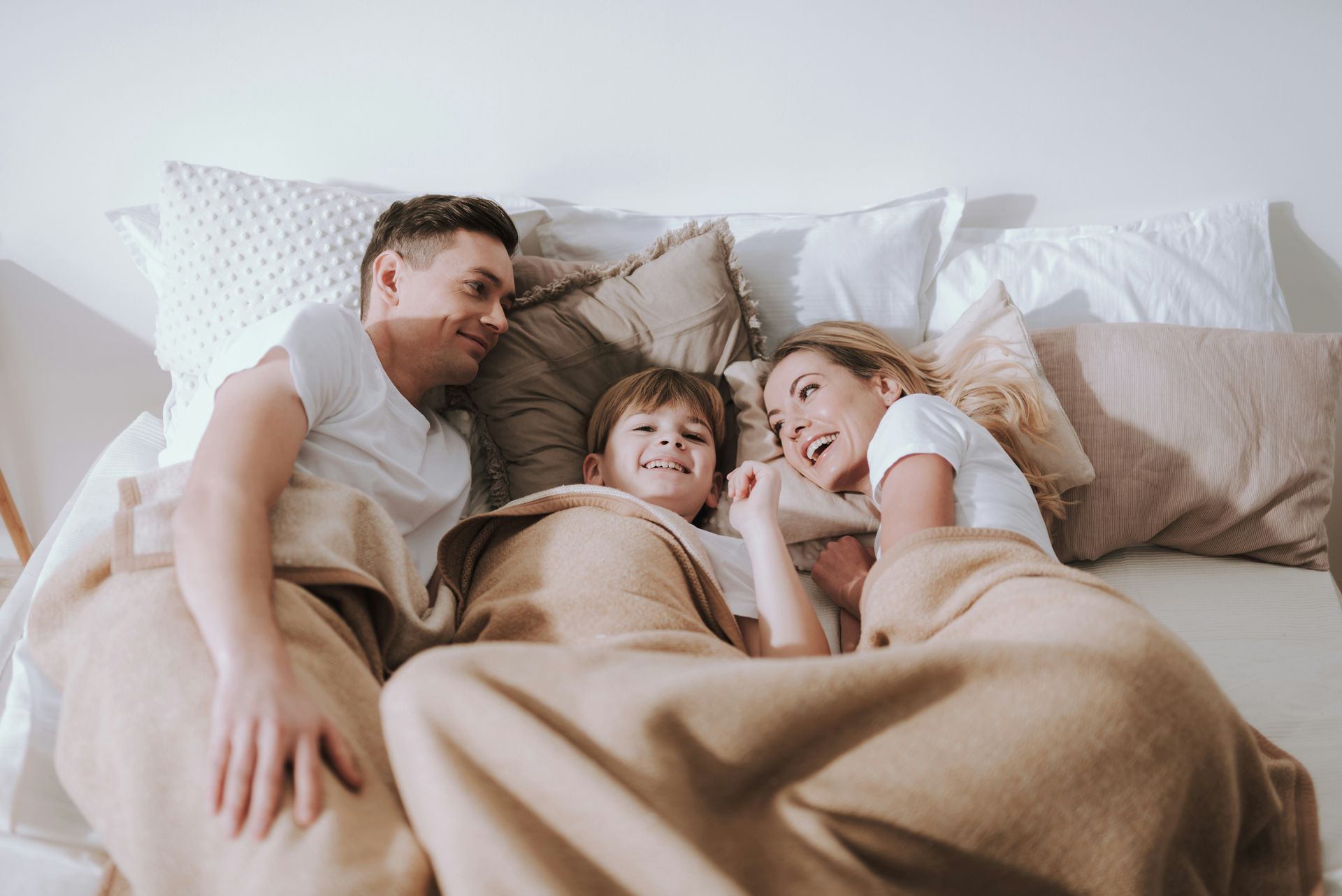 Family of three smiling, lying in bed under a blanket.