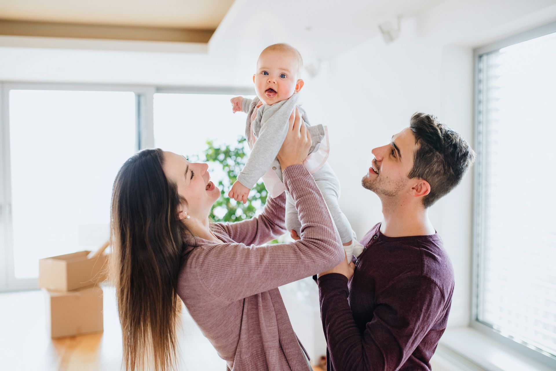 Parents joyfully lift a baby in a bright, empty room with moving boxes.