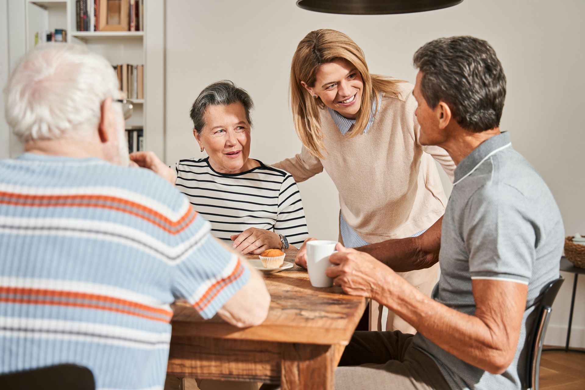 Four people at a table; woman smiles at two seated people while holding a mug. One person has striped shirt.